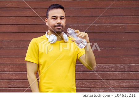 Brunet man with cordless earphones on neck in yellow t-shirt drinking water standing by wooden wall guy with open bottle walking and posing for camera outdoors 104613211