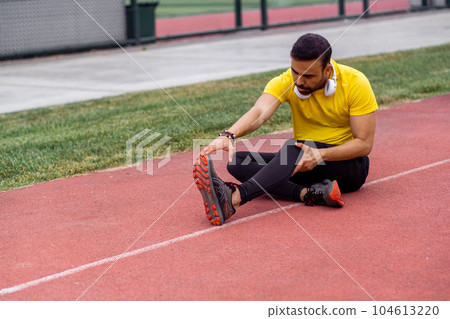 Focused athlete, wearing earphones, performs butterfly stretch on stadium track in a vibrant city park's urban sports arena. 104613220