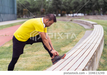 Man wearing yellow t-shirt tying sneaker laces leaning on bench with foot on sports ground in spring urban park runner man adjusting footgear at training 104613249