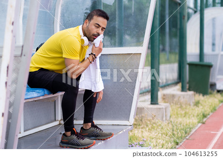 Fatigued man wiping sweat from face with clean towel sitting on chair in small arbor at urban sports stadium athlete resting after hard workout on outdoor athletic arena 104613255