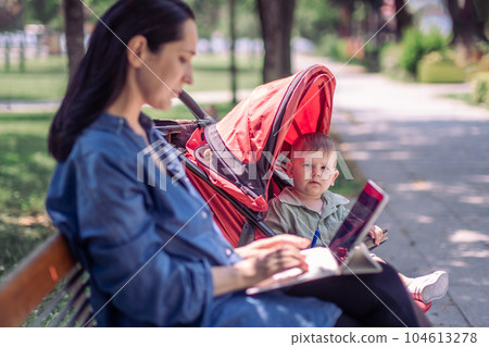 Curious little son looking at mother typing on laptop keyboard sitting in stroller in shady summer park woman freelancer with computer and baby boy in garden 104613278