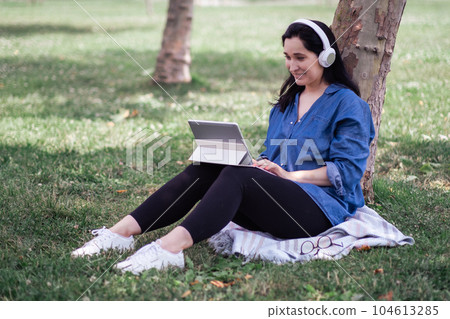 Smiling lady with headphones in denim shirt using video chat on tablet sitting on grass by tree in spring park woman listening to lecture on internet in garden Smiling lady with headphones in denim shirt using video chat on tablet sitting on grass by tree in spring park woman listening to lecture on internet in garden 104613285