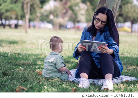 Business lady with smartphone and tablet sitting with little baby child in tree shadow in green park woman freelancer tries working online with son outdoors 104613313