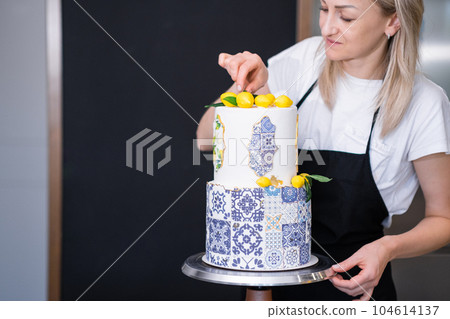 Focused housewife decorating large two-tiered cake with confit yellow lemons and green leaves cakes standing in kitchen blonde woman preparing custom confectionery for birthday party Focused housewife decorating large two-tiered cake with confit yellow lemons and green leaves cakes standing in kitchen blonde woman preparing custom confectionery for birthday party 104614137