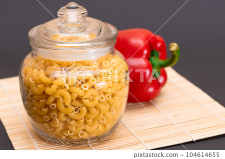 Uncooked Chifferi Rigati Pasta in Glass Jar with Red Bell Pepper on Bamboo Mat 104614655