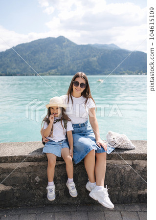Portrait of beautiful mom and daughter sitting on stone embankment. Background of big lake. Clear turquoise water 104615109