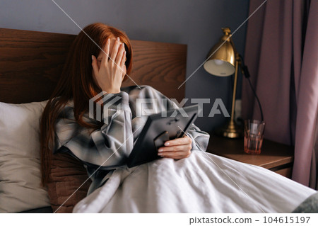 Side view of grief-stricken young woman lying on bed covering face with hand and crying while holding picture frame, touching photograph with love. Concept of nostalgia, grief, longing and loneliness. Side view of grief-stricken young woman lying on bed covering face with hand and crying while holding picture frame, touching photograph with love. Concept of nostalgia, grief, longing and loneliness. 104615197