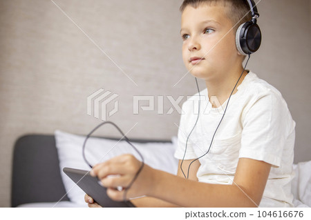 Smiling happy kid caucasian boy in T-shirt siting on bed, using tablet computer Smiling happy kid caucasian boy in T-shirt siting on bed, using tablet computer 104616676