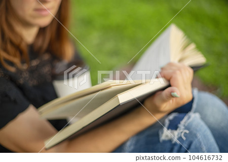 A woman sits near a tree in the park and holds a book and a cup with a hot drink 104616732
