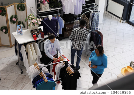 Top view of clients shopping for fashionable clothes in modern boutique, looking at hangers full with stylish merchandise, Diverse pepople checking new fashion collection, analyzing fabric 104619596