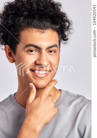 Portrait of a young handsome smiling guy with black curly hair displaying the results of consistent skincare and grooming routine in a studio. Swarthy man maintains his youthful and healthy appearance 104620543
