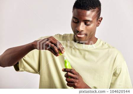 Black guy with youthful skin opens a bottle of facial oil or cream to apply it on his face. Handsome young African man maintaining his natural beauty with a skincare routine. Men's self-care. Black guy with youthful skin opens a bottle of facial oil or cream to apply it on his face. Handsome young African man maintaining his natural beauty with a skincare routine. Men's self-care. 104620544