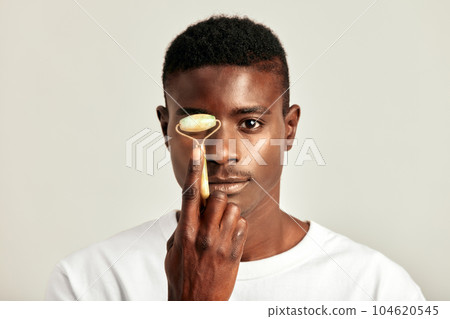 Black guy with youthful skin using jade de-puffing face roller to keep his skin soft and smooth. Handsome young African man maintaining his natural beauty with a skincare routine. Men's self-care. Black guy with youthful skin using jade de-puffing face roller to keep his skin soft and smooth. Handsome young African man maintaining his natural beauty with a skincare routine. Men's self-care. 104620545