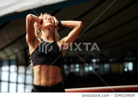 Young athlete girl getting ready for a workout at the stadium. Attractive slender blonde in a black tracksuit straightens and pins up her long hair before training. Active lifestyle and sports. 104620609