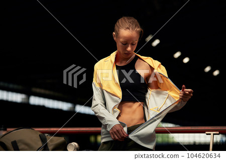 Young athlete girl getting ready for a workout at the stadium. Attractive slender blonde taking off a yellow-gray sports jacket before training. Active lifestyle, sports and fitness. Young athlete girl getting ready for a workout at the stadium. Attractive slender blonde taking off a yellow-gray sports jacket before training. Active lifestyle, sports and fitness. 104620634