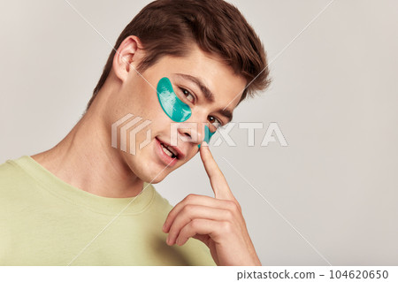 A millennial guy with brown hair practicing skincare with under eye hydrogel patches in a studio. Young smiling caucasian man applying cosmetic patches on his face. Men's self-care. A millennial guy with brown hair practicing skincare with under eye hydrogel patches in a studio. Young smiling caucasian man applying cosmetic patches on his face. Men's self-care. 104620650