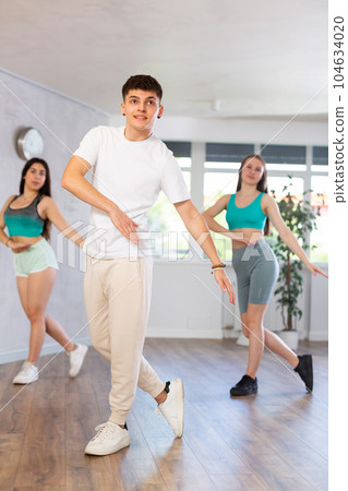 Guy and young girls in good mood actively move and dance reggaeton at choreography lesson Guy and young girls in good mood actively move and dance reggaeton at choreography lesson 104634020