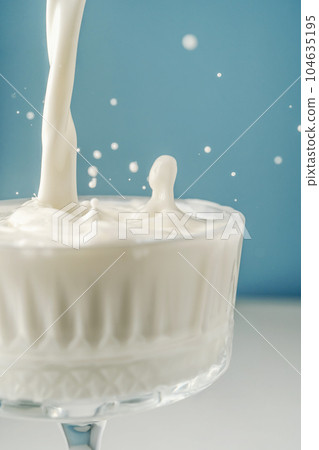 Milk pours into a beautiful crystal glass on a blue background and splashes around. Milk pours into a beautiful crystal glass on a blue background and splashes around. 104635195