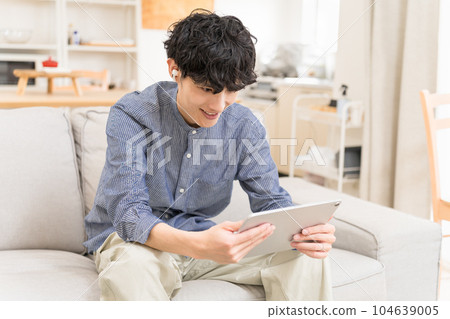 年輕人坐在沙發上，彎腰，戴著耳機，看著平板電腦，微笑 104639005