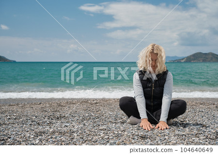 blonde european woman sits on a pebble beach on a spring day touches the stones on the beach with her hands. Outdoor portrait of a beautiful curly adult woman relaxing on the background of the sea blonde european woman sits on a pebble beach on a spring day touches the stones on the beach with her hands. Outdoor portrait of a beautiful curly adult woman relaxing on the background of the sea 104640649