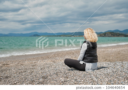 Beautiful adult blonde woman in sportswear sits on a pebble beach near the azure Aegean sea, natural sunlight, noon. View from the back, against the backdrop of sea and waves, rear view Beautiful adult blonde woman in sportswear sits on a pebble beach near the azure Aegean sea, natural sunlight, noon. View from the back, against the backdrop of sea and waves, rear view 104640654
