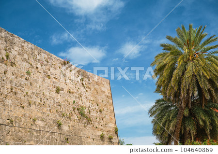 Fortress wall and palm trees on the street in the old shopping center of Rhodes, island of Rhodes, the Greek islands of the Dodecanese archipelago, holidays in Europe and popular tourist destination Fortress wall and palm trees on the street in the old shopping center of Rhodes, island of Rhodes, the Greek islands of the Dodecanese archipelago, holidays in Europe and popular tourist destination 104640869
