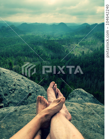 Crossed bare legs of a heterosexual couple cuddling on a cliff. Park background with rocks and forests. Crossed bare legs of a heterosexual couple cuddling on a cliff. Park background with rocks and forests. 104642240