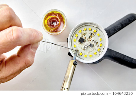 Soldering a burned-out LED into an LED light bulb. Hand with a soldering iron and rosin with solder on a white background Soldering a burned-out LED into an LED light bulb. Hand with a soldering iron and rosin with solder on a white background 104643230