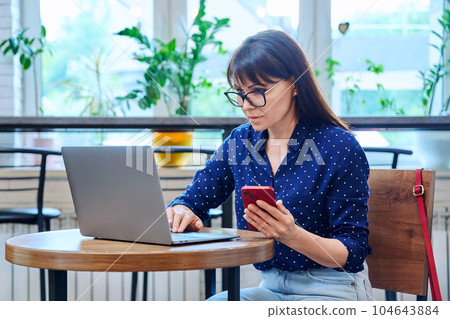 Middle-aged woman at coffee shop with smartphone laptop Middle-aged woman at coffee shop with smartphone laptop 104643884