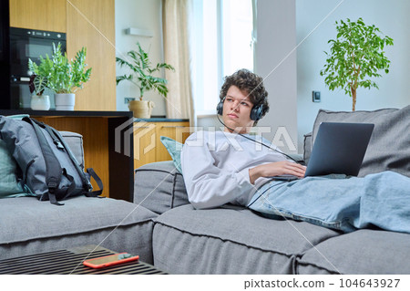 Young guy lying on sofa in living room, looking at laptop screen in headphones Young guy lying on sofa in living room, looking at laptop screen in headphones 104643927
