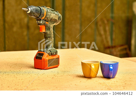 Electric drill,screwdriver on a plywood wooden surface. Cups, teatime, break Electric drill,screwdriver on a plywood wooden surface. Cups, teatime, break 104646412