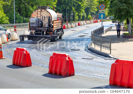 An industrial truck sprays liquid bitumen on a repaired section of a city road before laying new asphalt on a sunny day. An industrial truck sprays liquid bitumen on a repaired section of a city road before laying new asphalt on a sunny day. 104647170