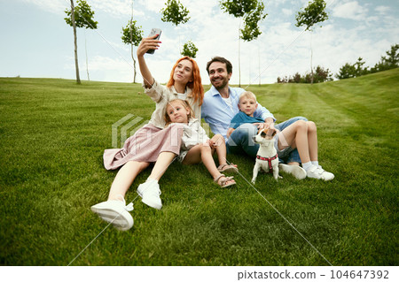 Young family, man and woman walking with their children in park, sitting on grass and taking selfie, photo together on warm summer day 104647392