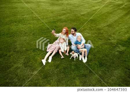 Young family, man and woman walking with their children in park, sitting on grass and taking selfie, photo together on warm summer day 104647393