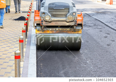 A metal cylinder of a road roller compacts fresh asphalt on a city street road on a sunny day. A metal cylinder of a road roller compacts fresh asphalt on a city street road on a sunny day. 104652303