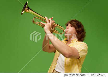 Portrait of young man with curly hair, talented musician playing trumpet against green studio background Portrait of young man with curly hair, talented musician playing trumpet against green studio background 104652323