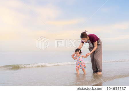 Asian mother holding hand daughter for support and walking on beach, Baby girl learning to walk, Happy family activity concept. 104655587