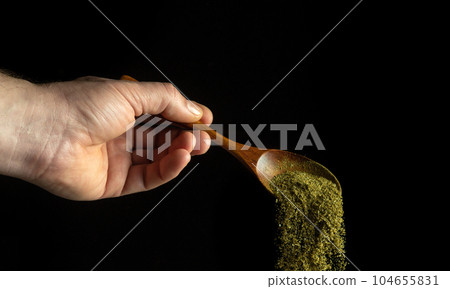 Mixture of dry chopped herbs is poured from a wooden spoon to add to a medicinal drink. Free space for advertising on a black background. 104655831