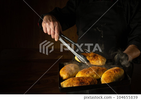 The baker bakes hot dog buns. Tongs in chef hand and baking sheet with hot pies. Place for advertising on a dark background 104655839