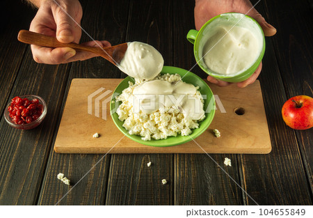 Close-up of a cook's hands with a spoon add milk yogurt to cheese on a kitchen black table while preparing a diet food 104655849