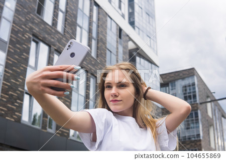 young woman taking a selfie or using a mirror app on the street against the backdrop of modern buildings 104655869