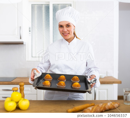 Cheerful female chef in white uniform holding baking tray of muffins Cheerful female chef in white uniform holding baking tray of muffins 104656337
