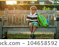 School boy sitting on bench with his backpack in park Primary school student after classes learning homework. Happy child in glasses waiting parents 104658592
