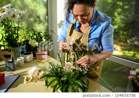 Florist inserts an orchid flower into a soaked foam inside a wicker basket while arranging bouquet with fresh flowers 104658786