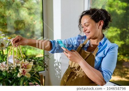 Smiling pregnant woman florist photographing a bouquet of orchid flowers in a wicker basket, sharing it on social media Smiling pregnant woman florist photographing a bouquet of orchid flowers in a wicker basket, sharing it on social media 104658796