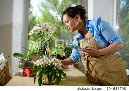 Lifestyle portrait. Female florist, entrepreneur in beige apron working with flower arrangements in floral design studio Lifestyle portrait. Female florist, entrepreneur in beige apron working with flower arrangements in floral design studio 104658798