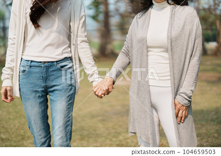 Adult daughter holding her elderly mother hand with love and walk together in park. Adult daughter holding her elderly mother hand with love and walk together in park. 104659503
