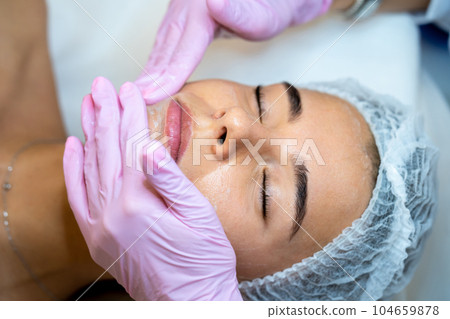 Unrecognizable female cosmetologist apply mask on woman's face, spa procedures, cleansing face. 104659878