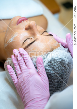 Woman cosmetologist apply cream on female's face, doing beauty procedures. 104659879