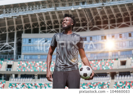 confident black football player holding the ball in his hand at the stadium 104660083